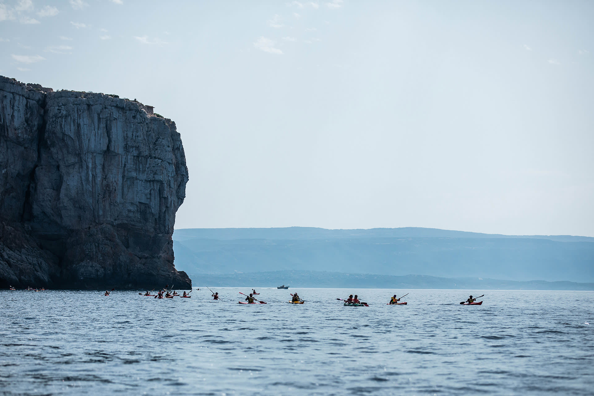A group of kayakers paddles on calm water near a large rocky cliff, with distant hills and a hazy sky in the background.