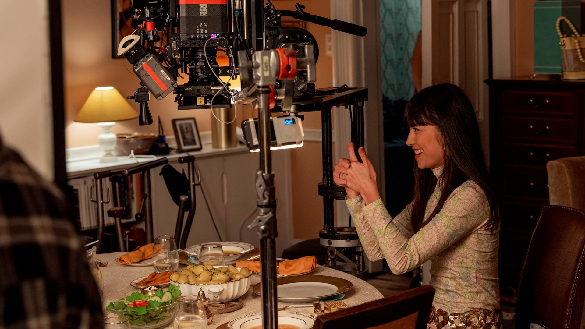 A woman sits at a dining table set for a meal, smiling and gesturing with her hands, while a film camera and crew equipment are positioned in front of her during a scene shoot inside a warmly lit room.