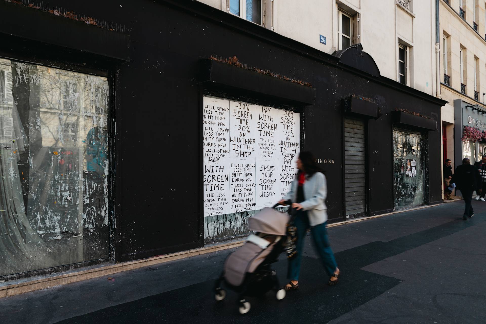 A person pushes a stroller past a black-painted building with posters on the windows. The posters repeatedly state, “PAY WITH TIME,” “SHARE THE PAIN,” and similar phrases in black text on white paper.