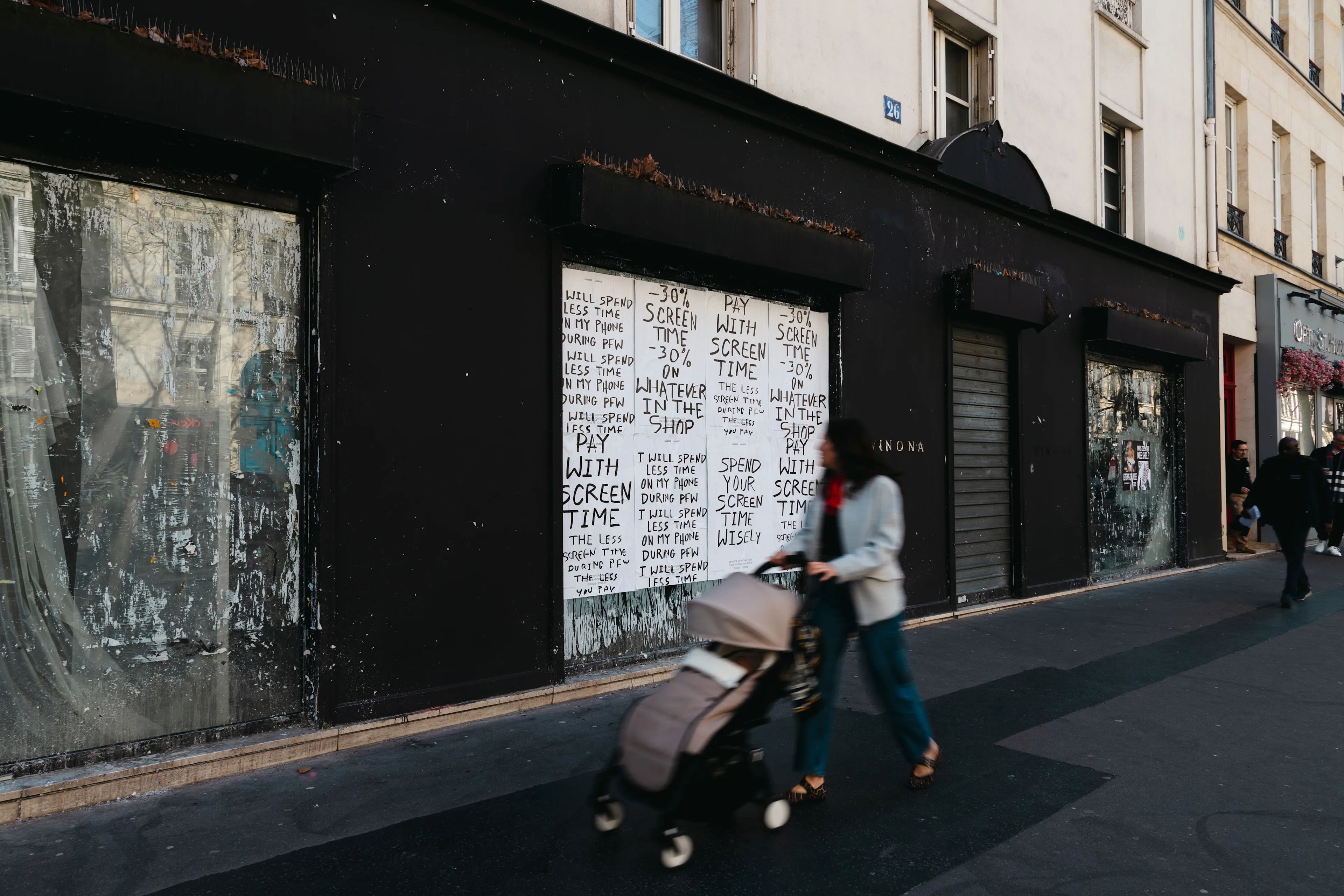A person pushes a stroller past a black-painted building with posters on the windows. The posters repeatedly state, “PAY WITH TIME,” “SHARE THE PAIN,” and similar phrases in black text on white paper.
