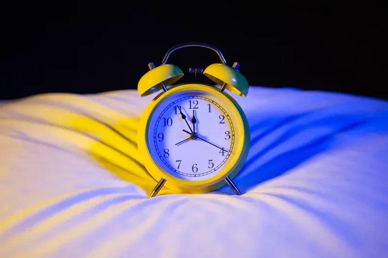 A yellow alarm clock showing 10:09 sits on a white pillow, with blue and yellow lighting creating shadows on the bedding.