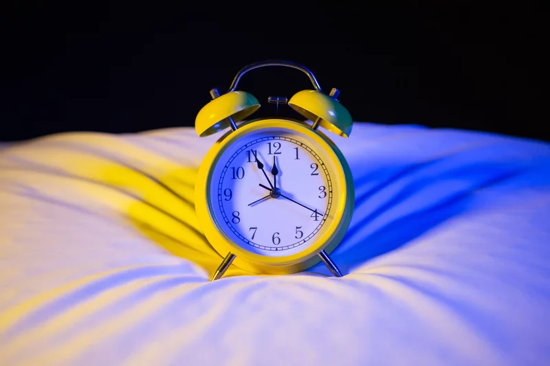 A yellow alarm clock showing 10:09 sits on a white pillow, with blue and yellow lighting creating shadows on the bedding.