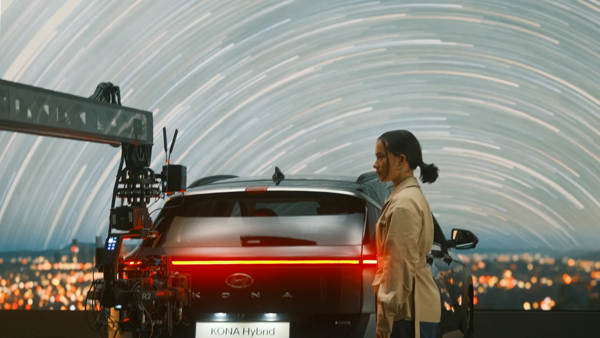 A woman stands beside a Hyundai Kona Hybrid, with a camera rig filming and a backdrop of swirling star trails and city lights behind them.