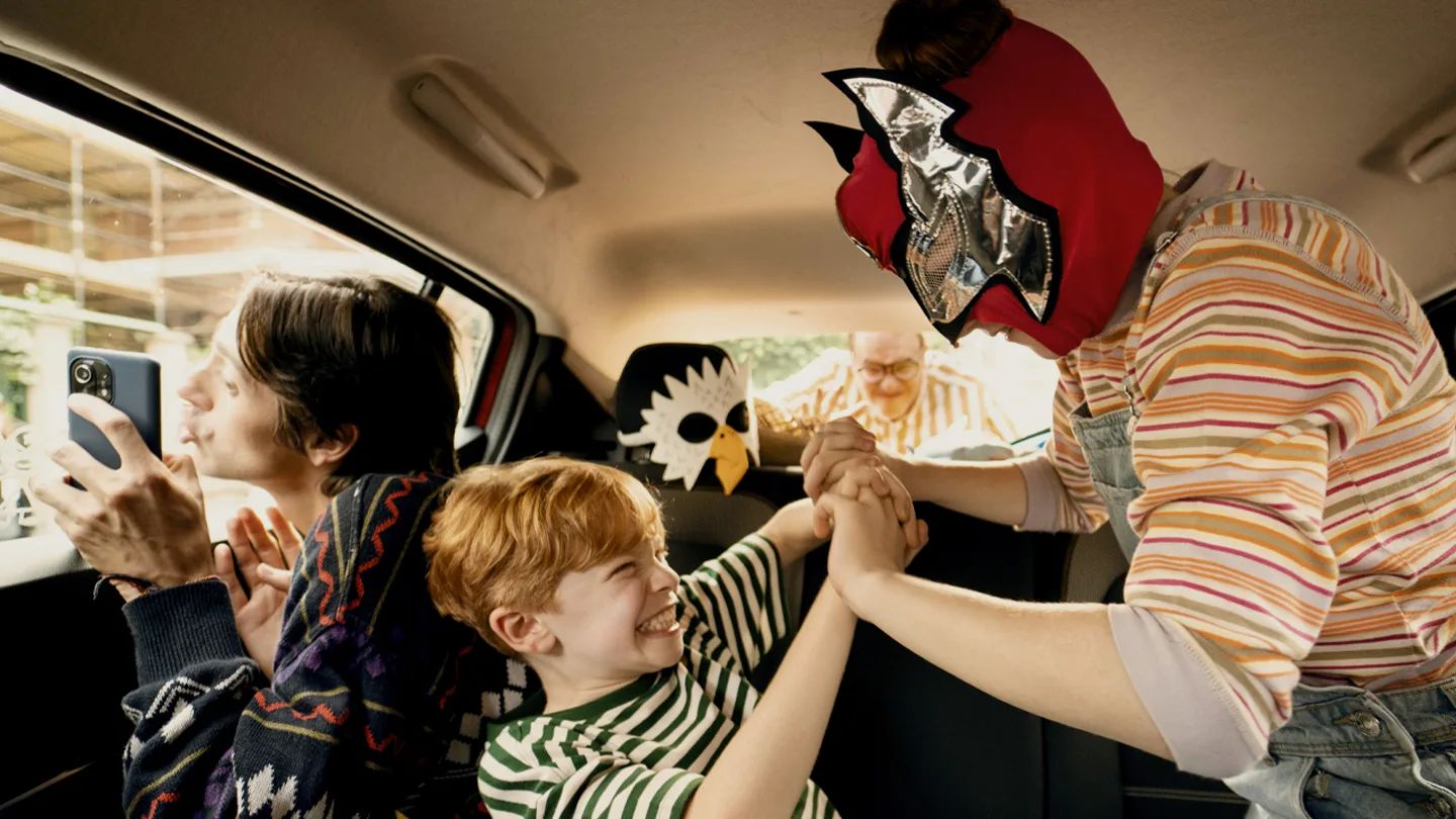 A child in a striped shirt laughs while being playfully wrestled by another child wearing a red and silver mask inside a car, as an adult next to them smiles and takes a photo with a phone.