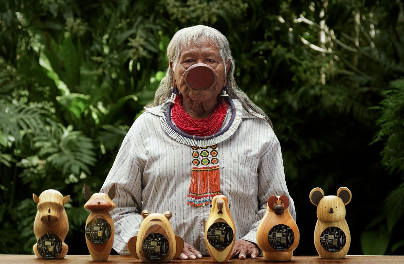 An Indigenous woman with long gray hair and a large lip plate stands behind a table displaying six animal-shaped wooden figurines, set against a lush green background. She wears traditional jewelry and clothing.