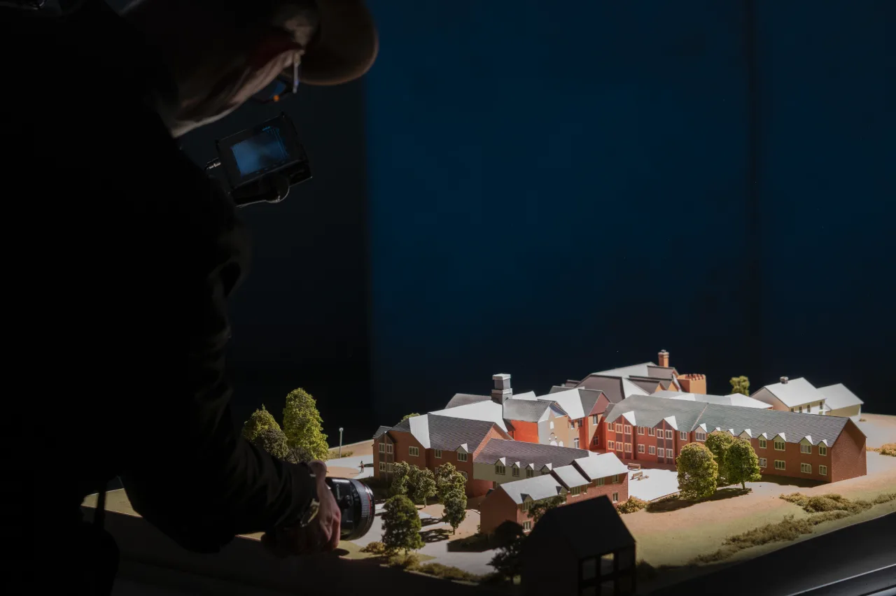 A person takes photos of a detailed architectural model of a building complex with trees, under dramatic lighting that highlights the model’s features.