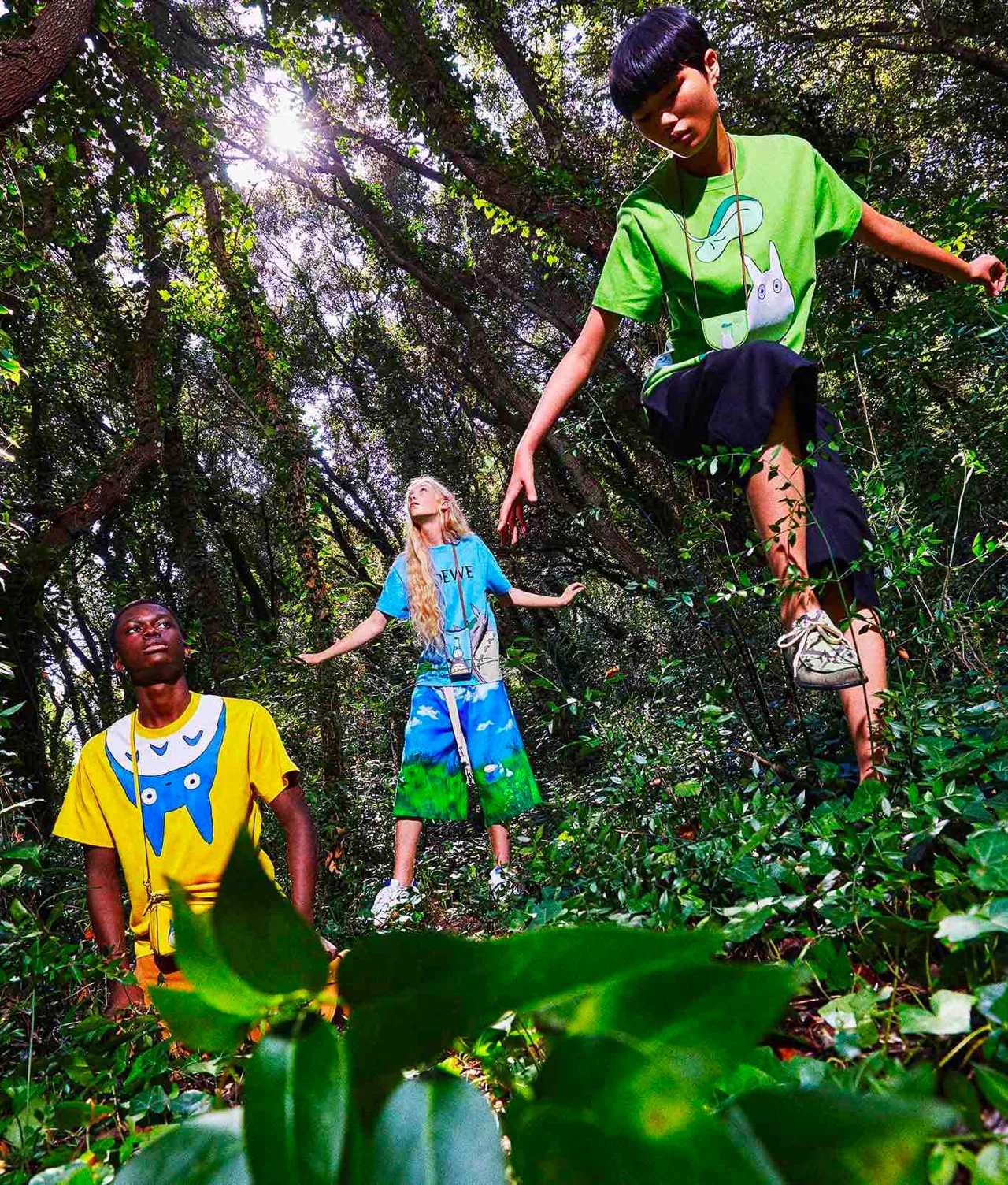 Three people stand in a sunlit, leafy forest wearing colorful Totoro-themed shirts and shorts. Sunlight filters through the trees above, creating a mystical, vibrant atmosphere.