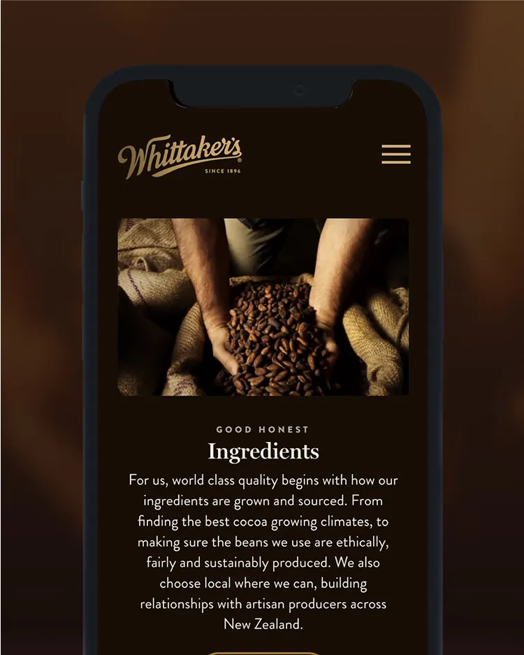 A smartphone screen displays Whittaker’s branding and a photo of hands holding cocoa beans over a pile, with text about sourcing ethical and local ingredients for chocolate.