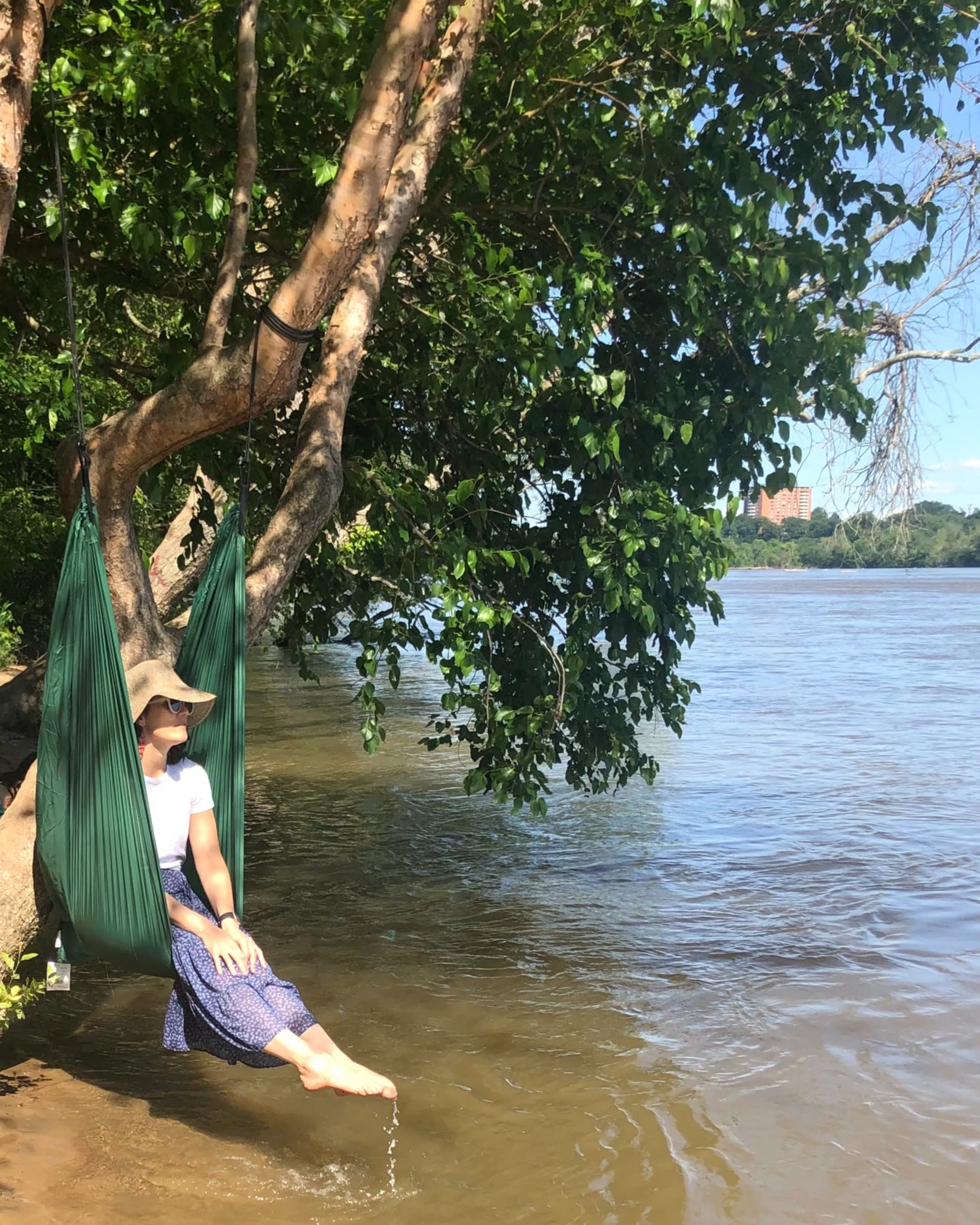 A woman in a sunhat sits in a green hammock tied to a tree by the water’s edge, with her feet gently touching the river. Lush green foliage hangs overhead, and sunlight reflects off the calm water.