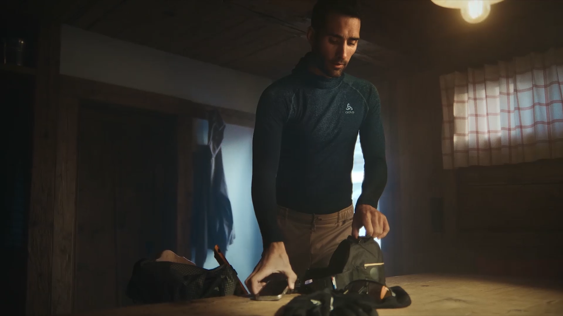 A man in a dark long-sleeve shirt stands at a wooden table in a dimly lit room, packing or organizing items, with warm light coming from above and a window with a checked curtain in the background.
