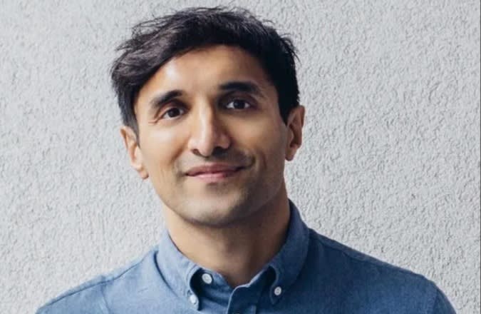 A man with short dark hair, wearing a blue button-up shirt, stands in front of a textured light gray wall, smiling gently at the camera.