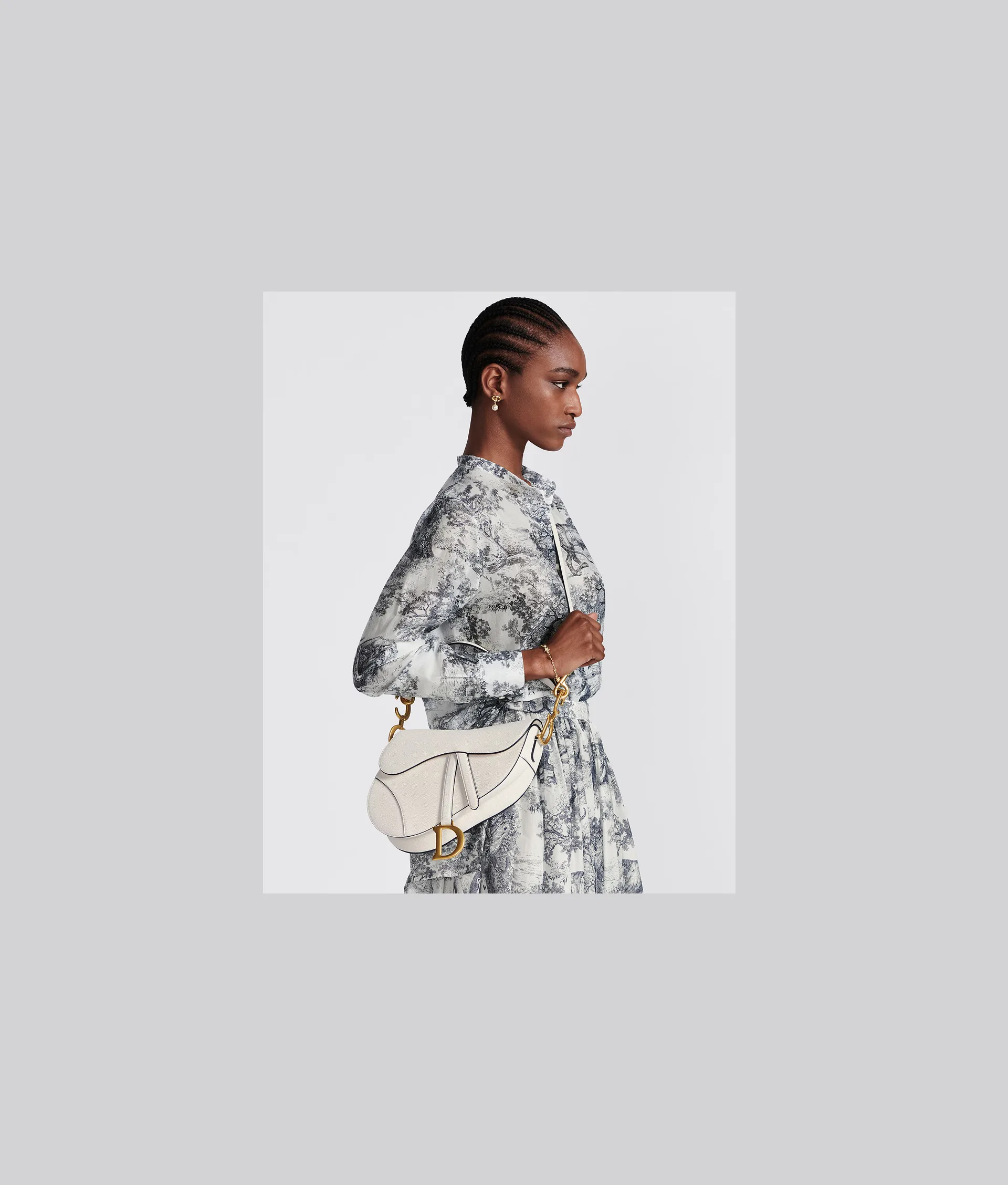 A woman with braided hair wears a long-sleeve, black-and-white patterned dress and holds a white designer handbag with a gold clasp, standing in profile against a light gray background.
