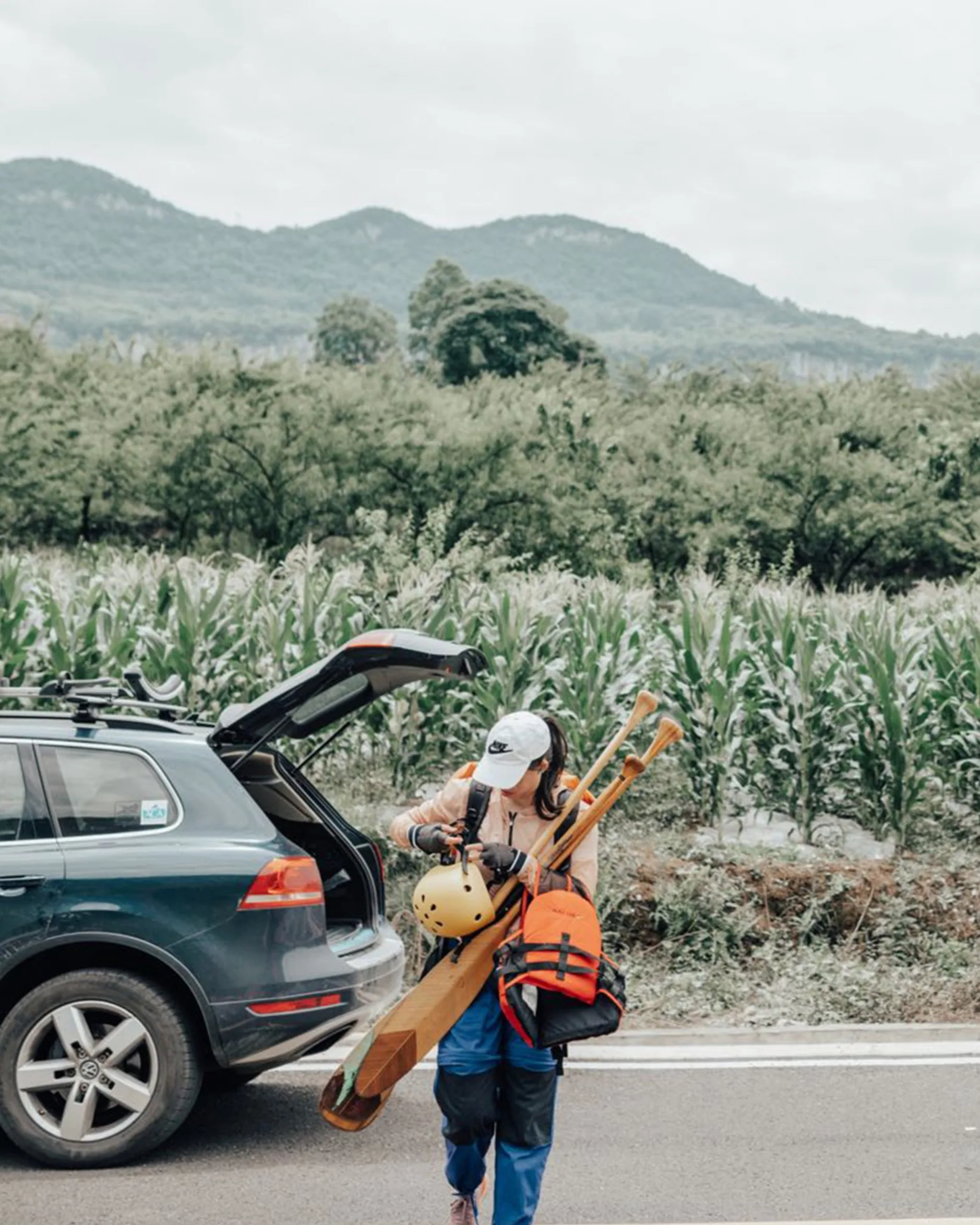 A person wearing a cap carries paddles, a helmet, and a life jacket while walking away from an open car trunk, with green fields and mountains in the background.