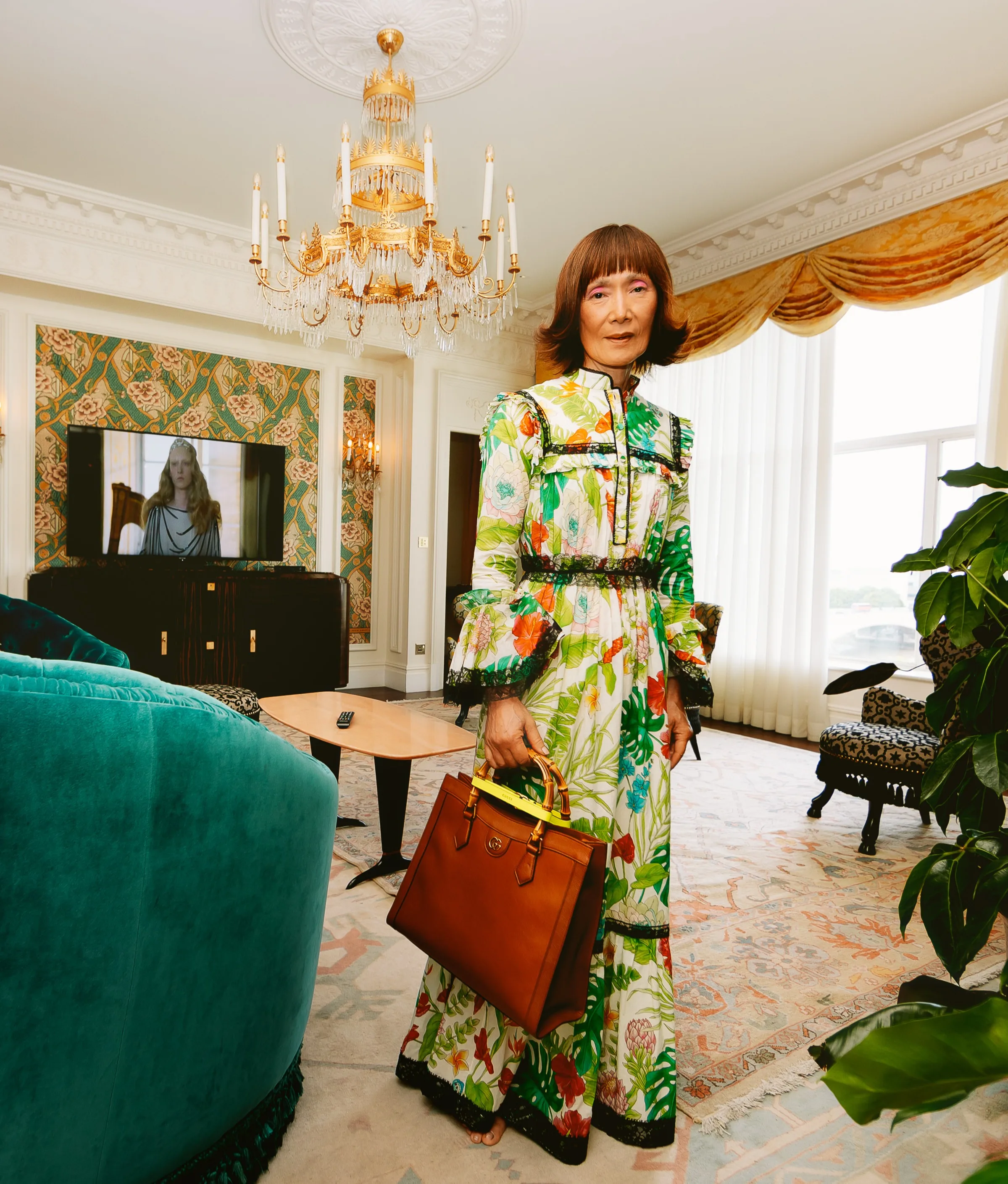 A woman in a colorful floral dress stands in a luxurious, elegant living room with ornate decor, a large chandelier, and a turquoise sofa, holding a brown handbag. A TV in the background displays a scene with a woman in white.