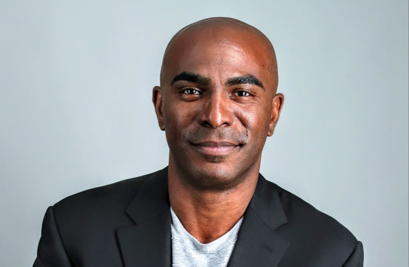 A man with a shaved head and dark complexion wearing a black blazer over a gray t-shirt, smiling slightly at the camera against a plain light gray background.