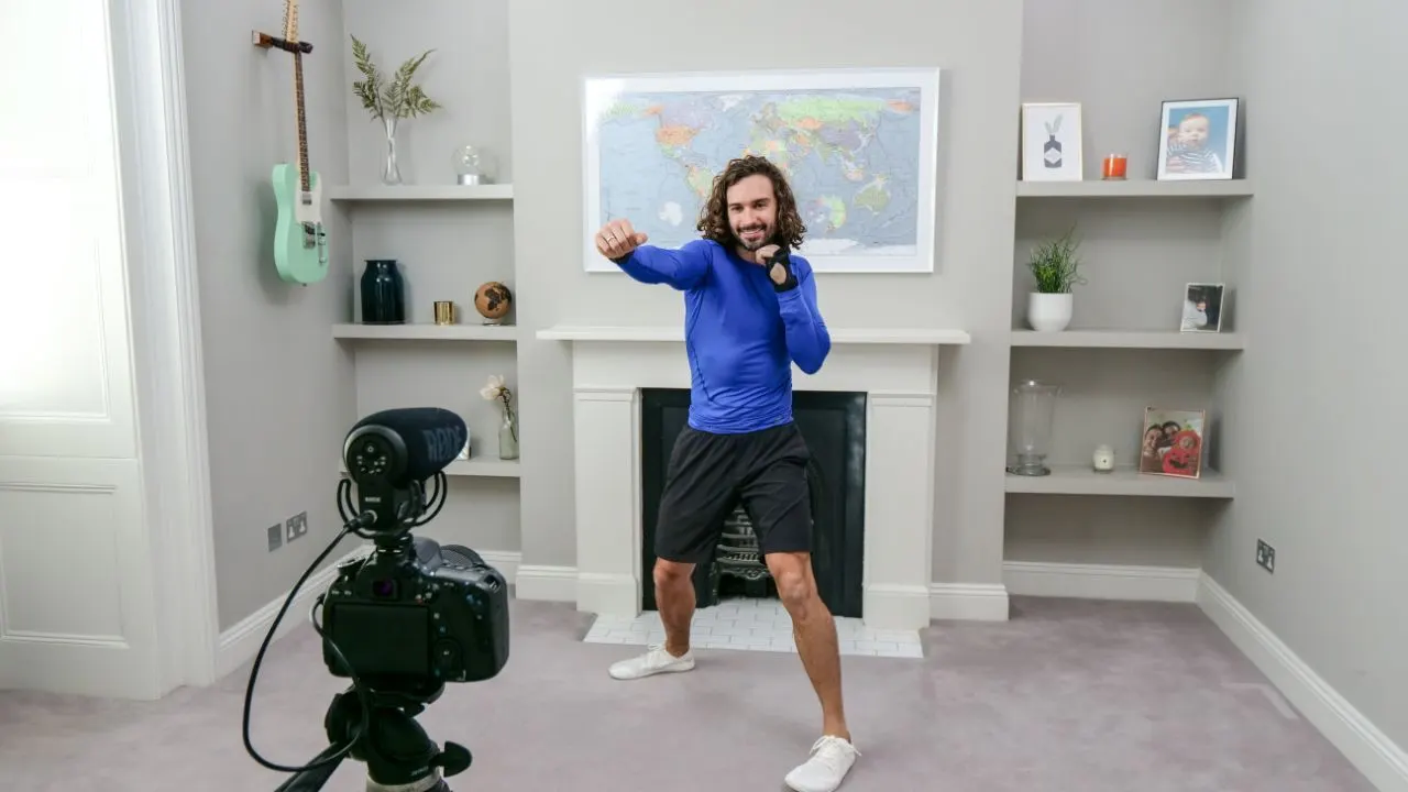 A man in sportswear exercises in his living room in front of a camera, punching the air with both fists. Shelves with photos and decorations, a green guitar, and a world map are visible in the background.