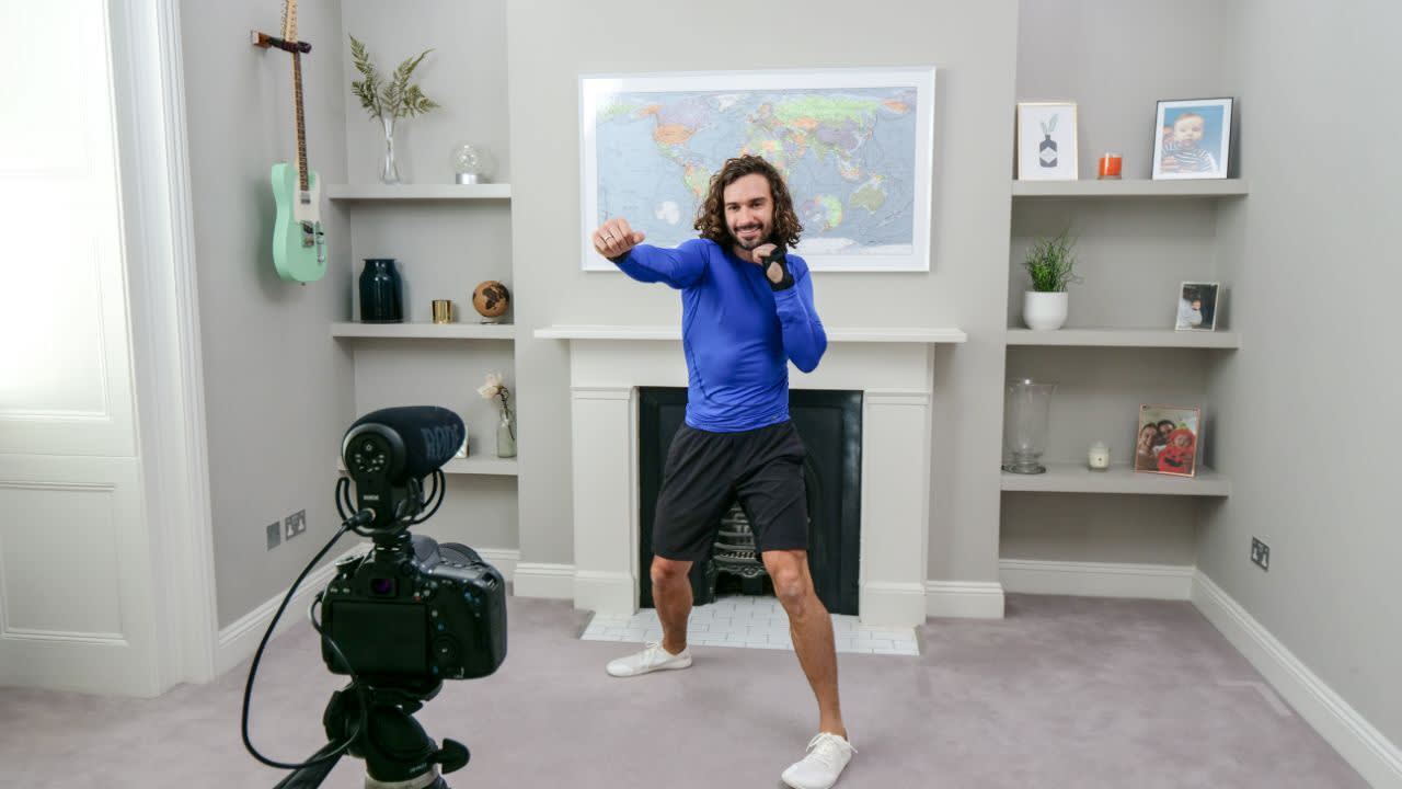 A man in sportswear exercises in his living room in front of a camera, punching the air with both fists. Shelves with photos and decorations, a green guitar, and a world map are visible in the background.