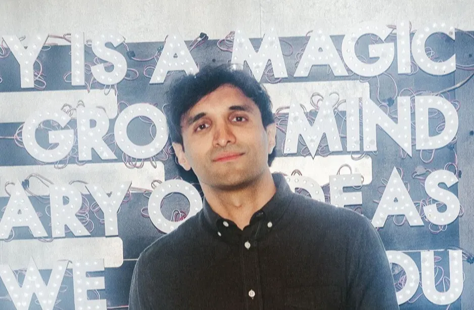 A man with dark hair wearing a dark button-up shirt stands in front of a wall with large, partially obscured, white text and decorative lights.