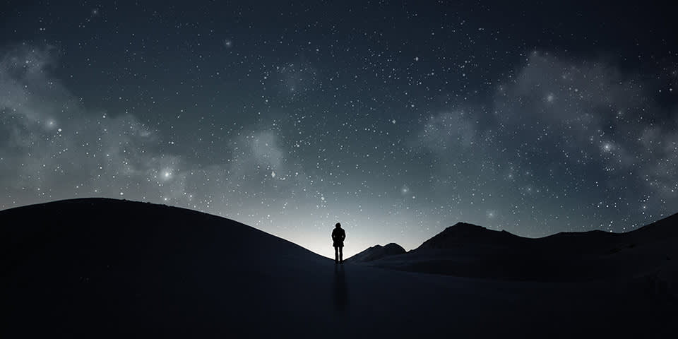 A lone person stands on a hill under a dark, starry night sky with scattered clouds, surrounded by silhouettes of distant mountains.