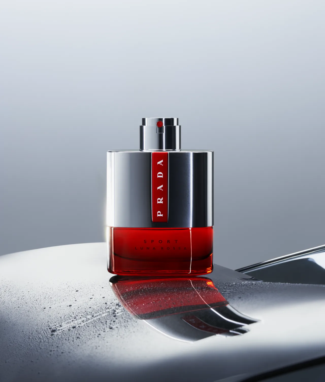 A bottle of Prada Luna Rossa Sport perfume with a red base and silver top sits on a glossy surface, reflecting its image.