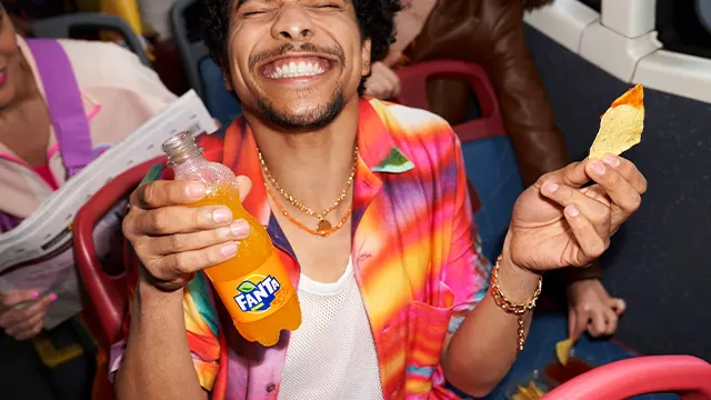 A smiling man in a colorful shirt holds an orange Fanta bottle and a potato chip with dip, sitting on a bus with friends. Another person nearby is reading a newspaper. The scene is cheerful and lively.