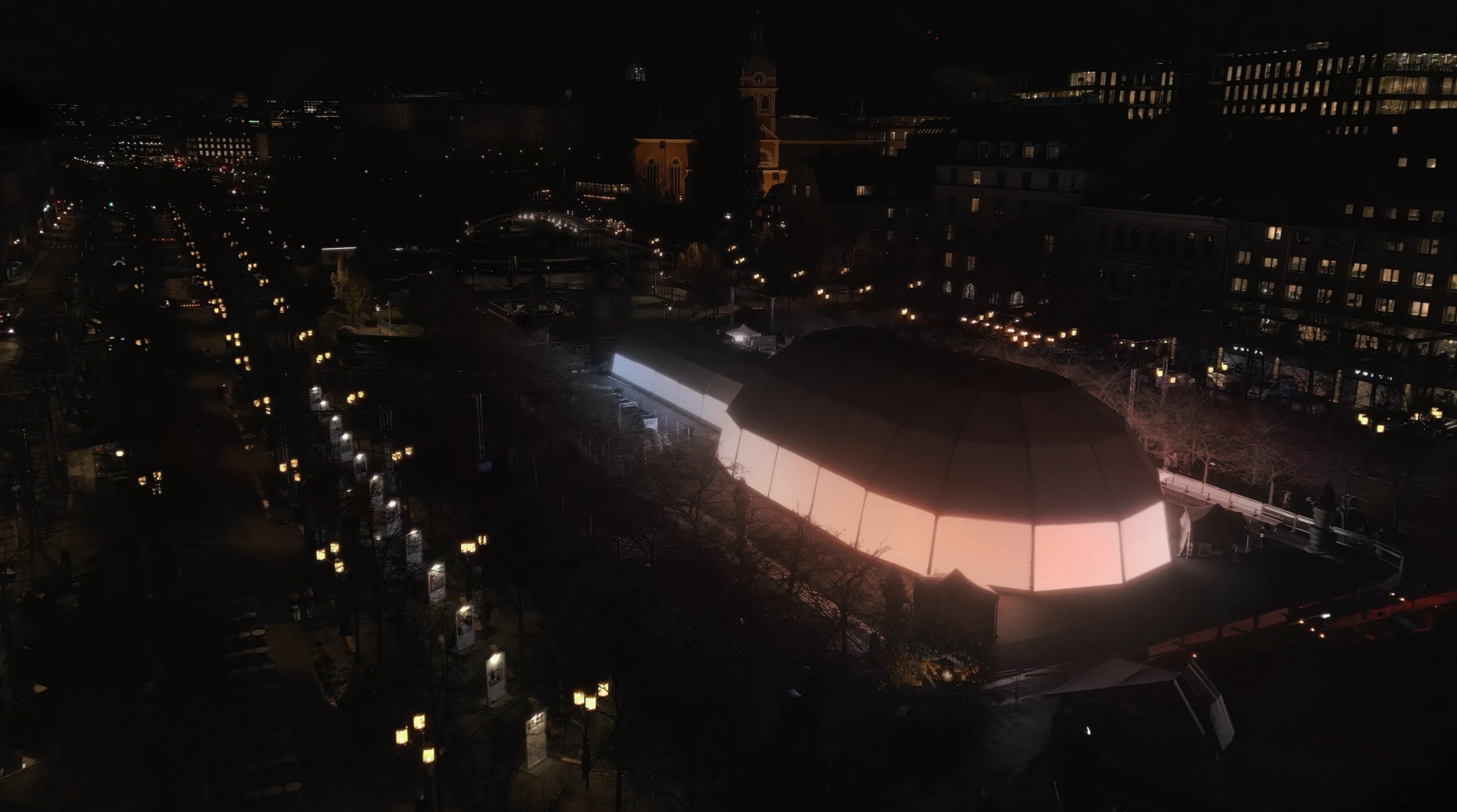 A large, dome-shaped building glows with soft pink and white lights at night, surrounded by a cityscape with illuminated streets and historic buildings in the background.