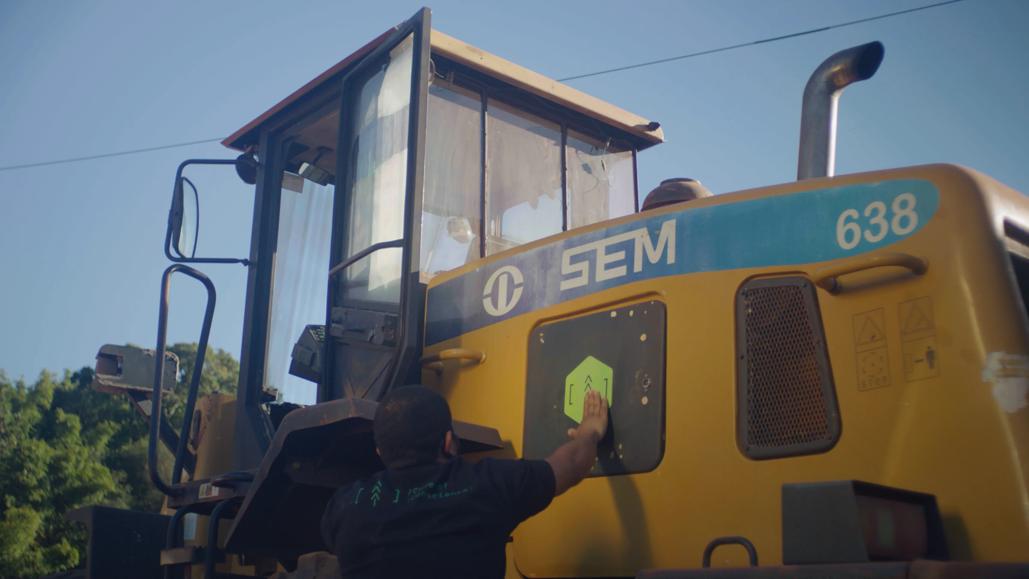 A person places a green sticker on the side of a large yellow construction vehicle marked SEM 638 under a clear blue sky. Trees are visible in the background.