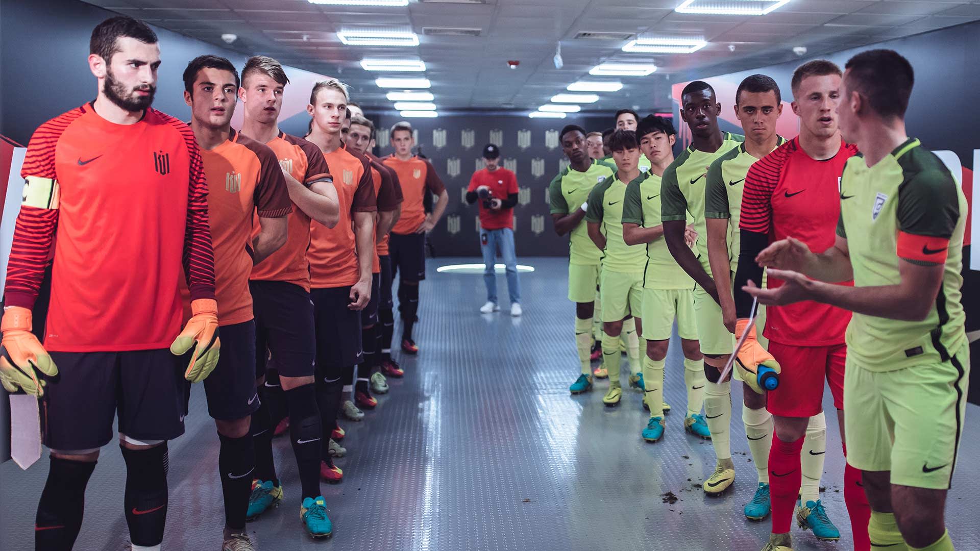 Two soccer teams stand facing each other in a hallway before a match. One team wears orange and black kits, the other light green kits. The players look focused, with some talking amongst themselves.