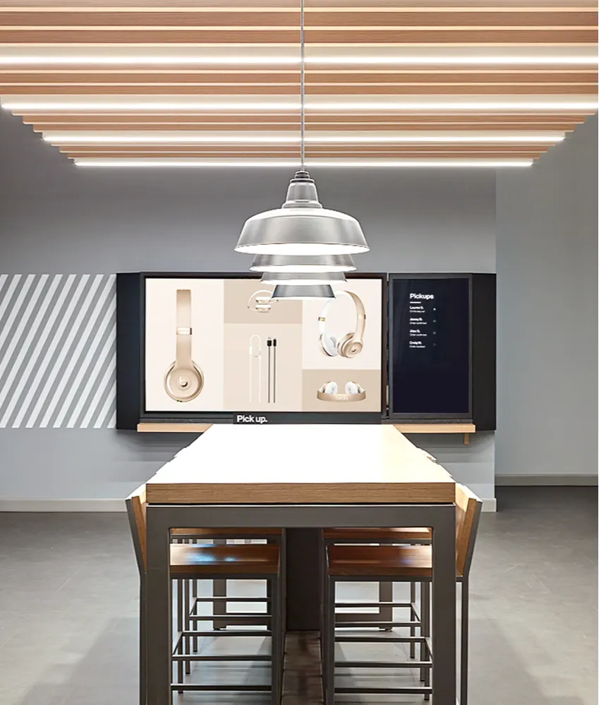 A modern store interior with a long wooden table and stools, pendant lighting above, and digital screens on the wall displaying images of headphones, earbuds, and a “Pick up” notice.