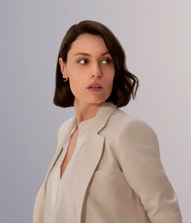 A woman with short brown hair wearing a cream blazer and white blouse looks to her left against a light, neutral background.