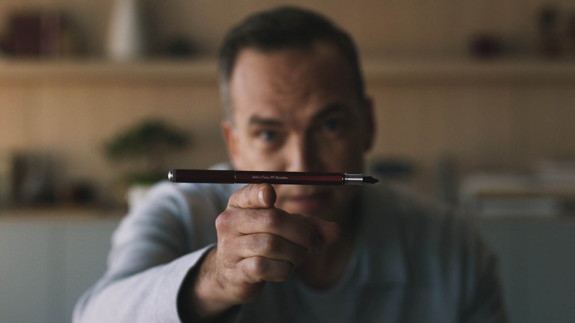 A person in focus holds a red pen pointed toward the camera, with the pen in sharp focus and the persons face blurred in the background. The setting appears to be indoors.
