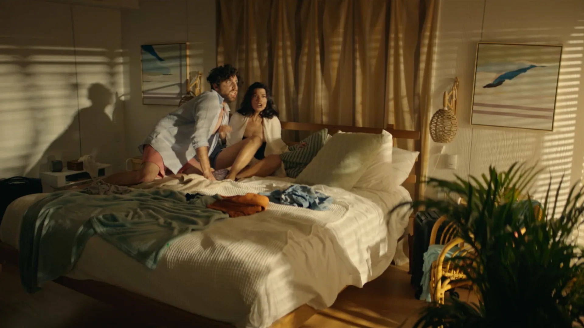 A man and woman sit surprised on a messy bed in a sunlit bedroom, partially covered by a blanket. Clothes are scattered around, and the room is decorated with paintings and plants.