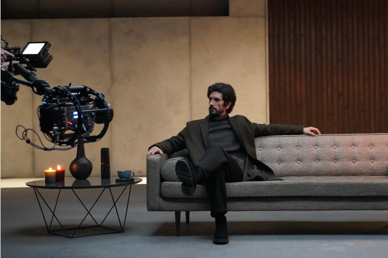 A man with dark hair and a beard sits on a gray sofa in a modern, minimalistic room. A camera is filming him. A lit candle, cup, and book are on a nearby glass coffee table. The background features concrete and wooden walls.