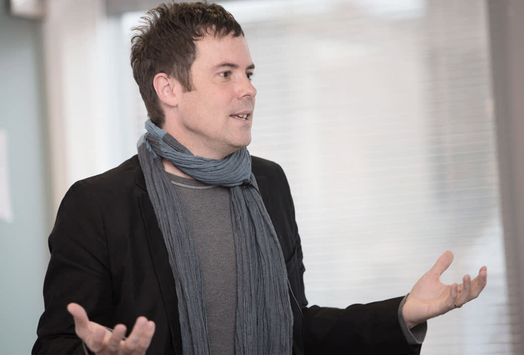 A person with short dark hair, wearing a dark jacket, grey shirt, and a blue scarf, stands indoors with their hands raised, as if speaking or explaining something. The background is softly blurred.