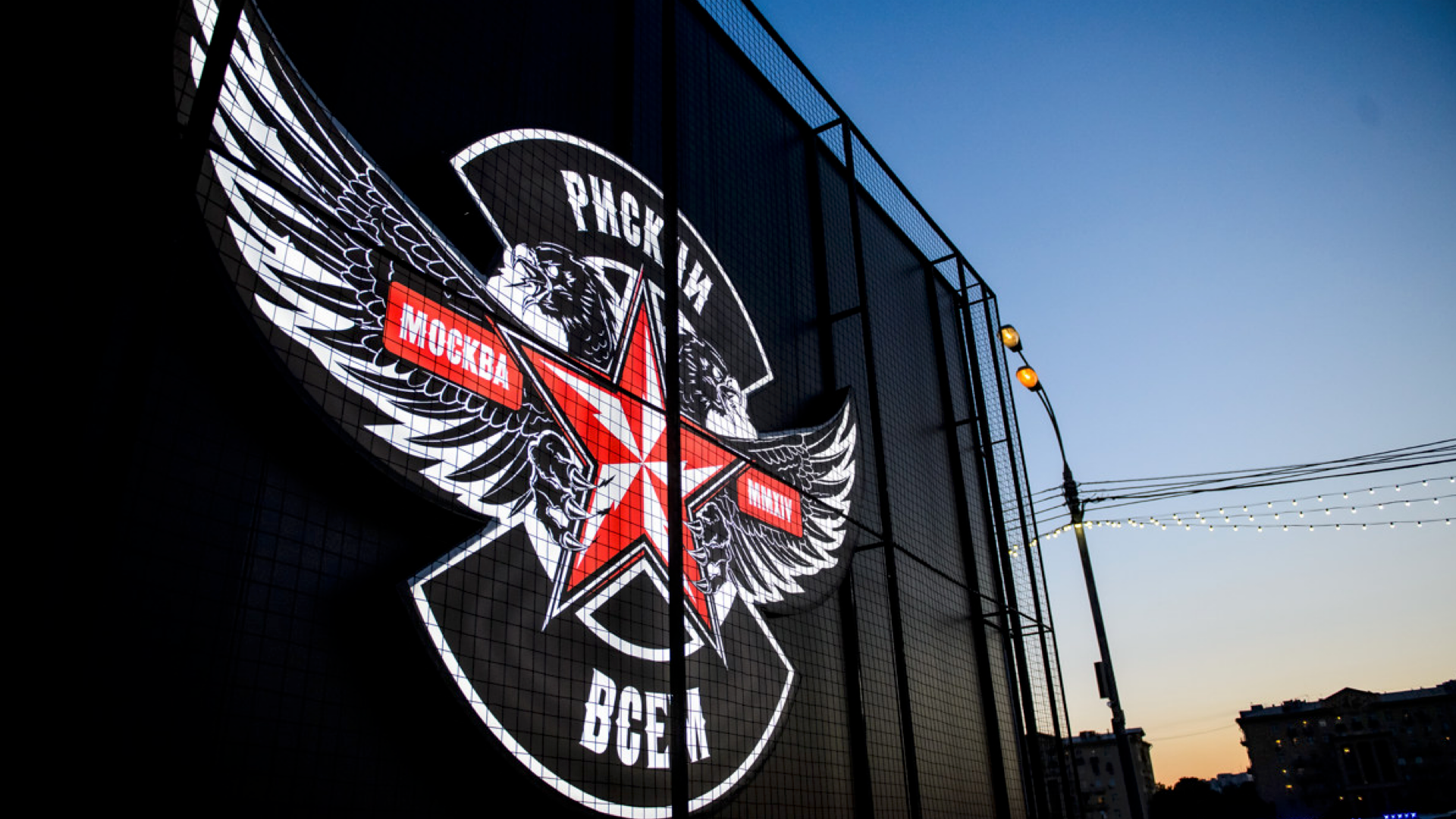 Large illuminated sign on a building features a red star with wings, Russian text, and two lion heads. The evening sky and city streetlights are visible in the background.