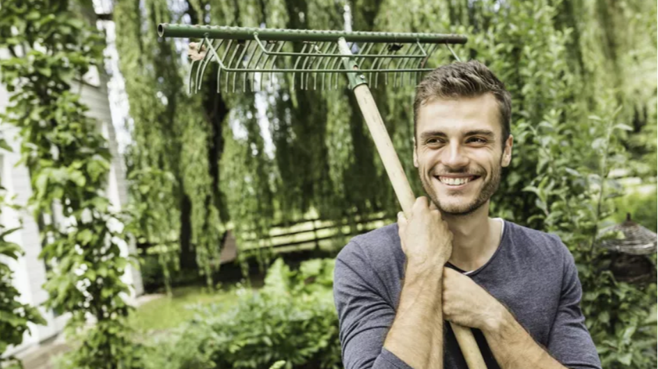A smiling man stands outdoors holding a garden rake over his shoulder, with green trees and plants in the background on a sunny day.