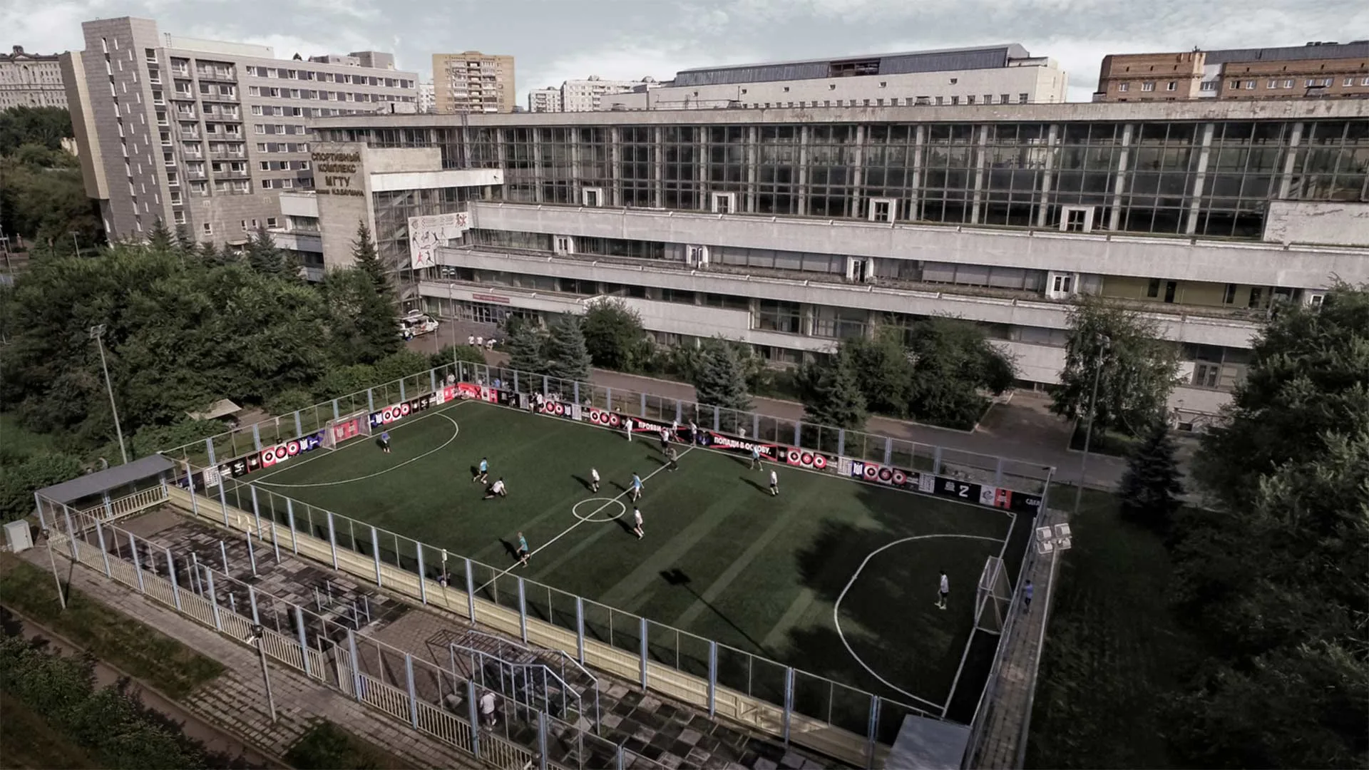 Aerial view of a small outdoor soccer field with players in action, surrounded by a metal fence, located next to a large, multi-story building in an urban area with trees and other buildings nearby.