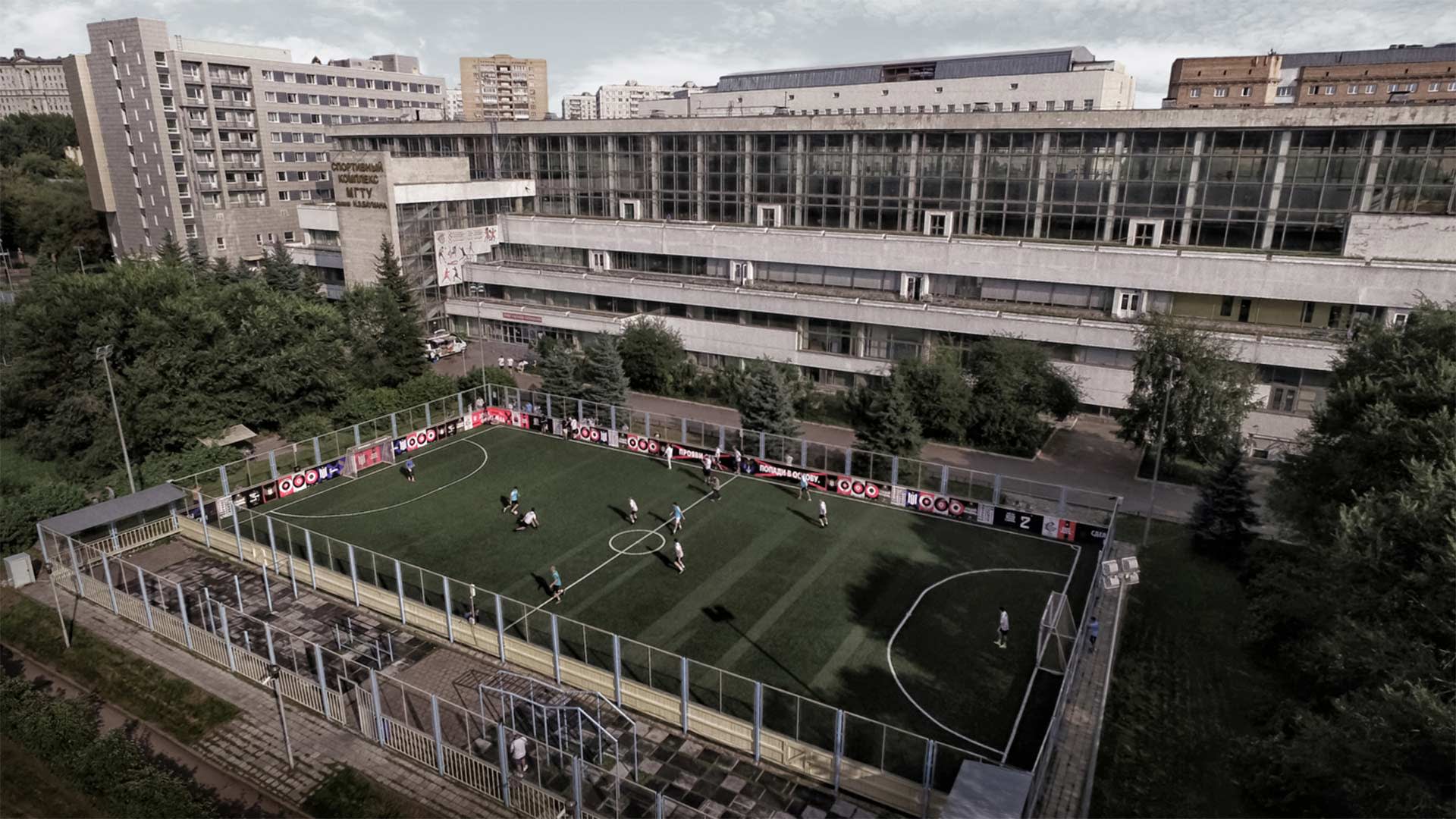 Aerial view of a small outdoor soccer field with players in action, surrounded by a metal fence, located next to a large, multi-story building in an urban area with trees and other buildings nearby.