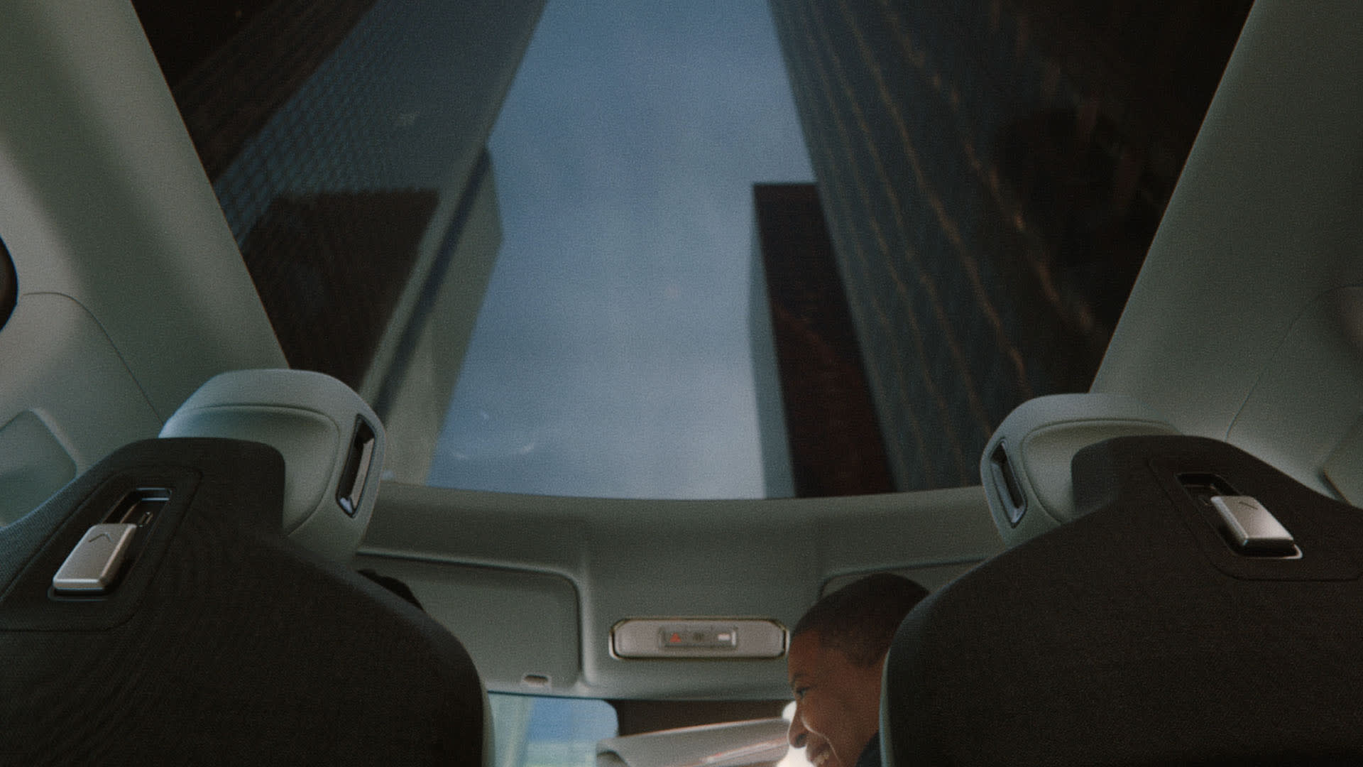 View from inside a car with a glass roof, looking up at tall city buildings. Two front seats are visible, with a person partially seen in the passenger seat, and a blue sky above.