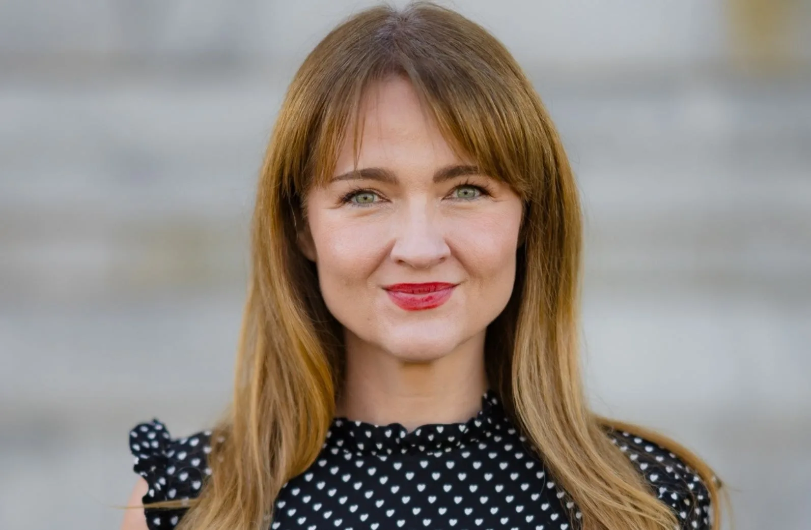 A woman with straight, light brown hair and green eyes smiles at the camera. She is wearing a black top with small white heart patterns. The background is blurred and gray.
