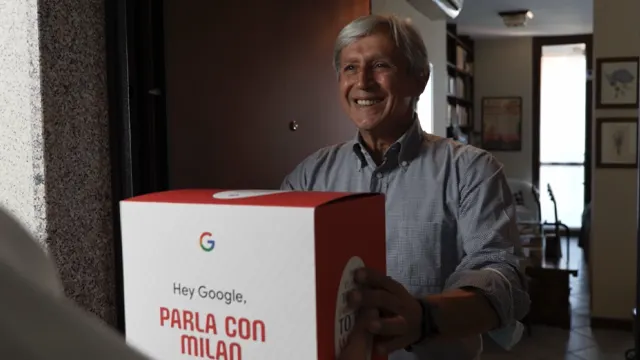 An older man with gray hair smiles as he receives a large Google-branded box at his door. The box has Italian text that starts with “Hey Google, PARLA CON MILAO.” The scene is indoors with natural light.