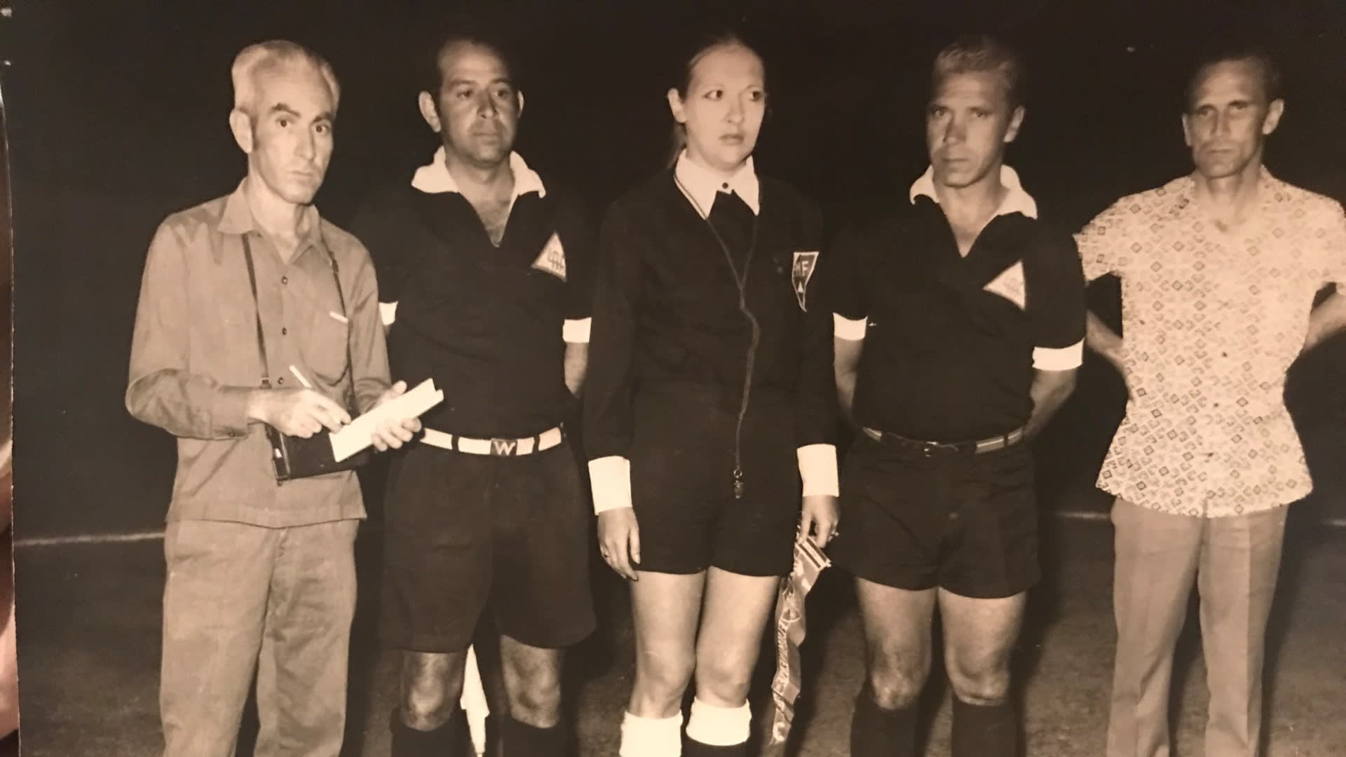 A black-and-white photo of five adults standing side by side. Three are dressed as soccer referees, one holds a notebook, and another, in a light-patterned shirt, stands at the far right. They appear to be on a soccer field at night.