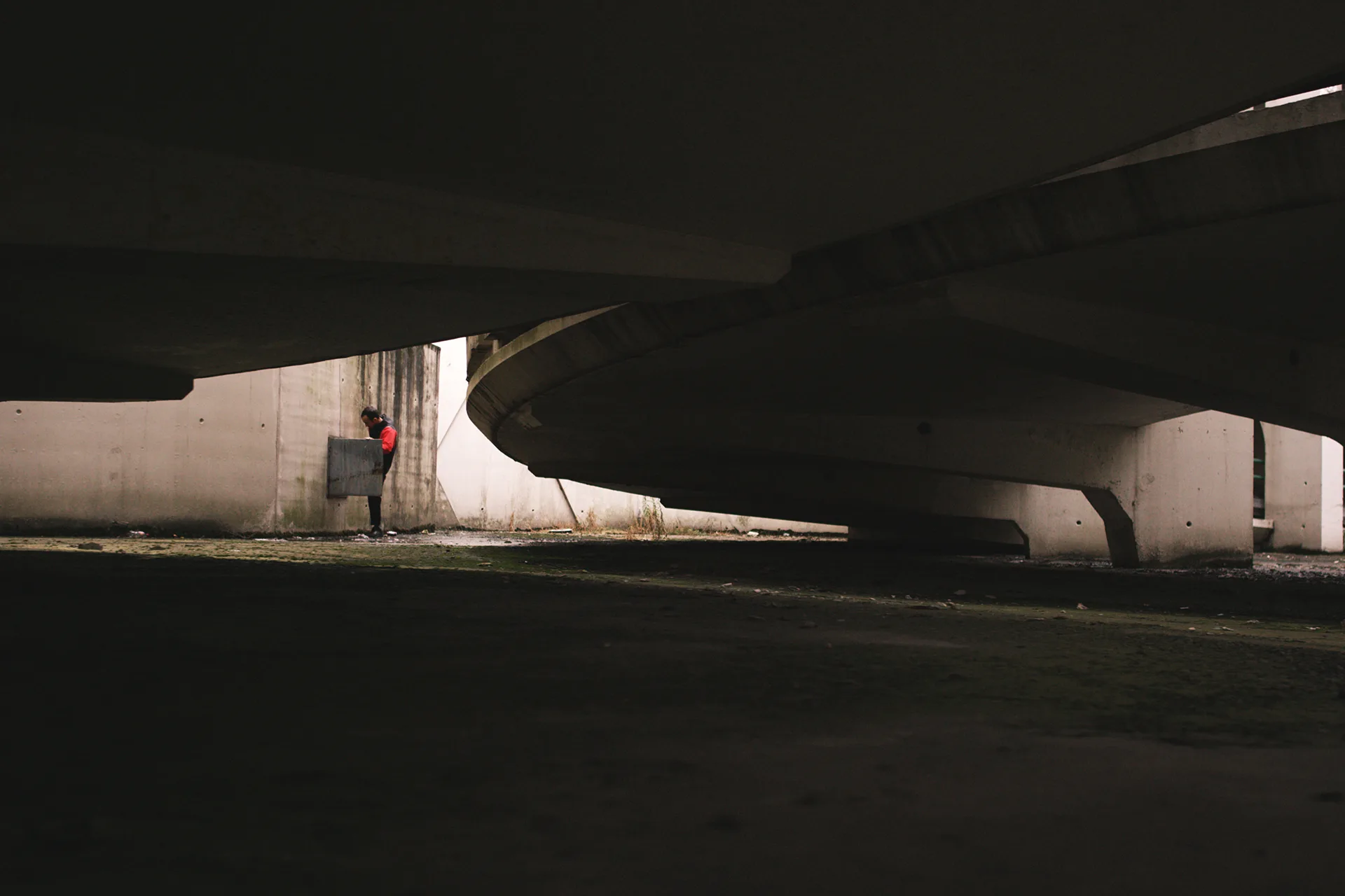 A person in a red shirt stands against a concrete wall in a dimly lit, industrial space beneath large, curved, overhead structures. The foreground is dark with patches of moss or dirt on the ground.