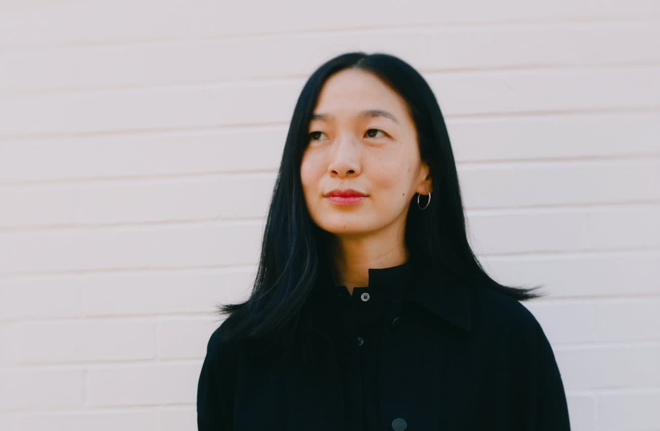 A woman with long black hair wearing a black coat and gold hoop earrings stands in front of a white brick wall, looking slightly to the side with a calm expression.