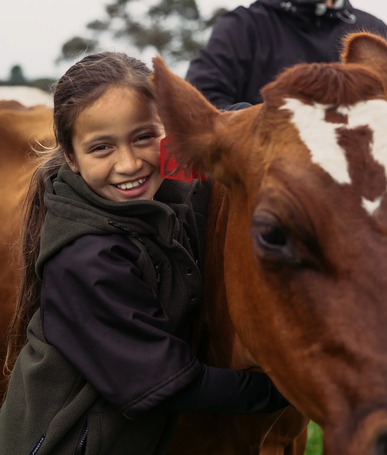 A smiling girl with long brown hair hugs a brown cow with a white mark on its forehead, standing outdoors. Another person in dark clothing is partially visible in the background.