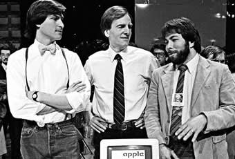 Three men in business attire stand around an early Apple computer. The man on the left wears a bow tie and suspenders, while the other two wear suits and ties, engaging in conversation at a tech event.