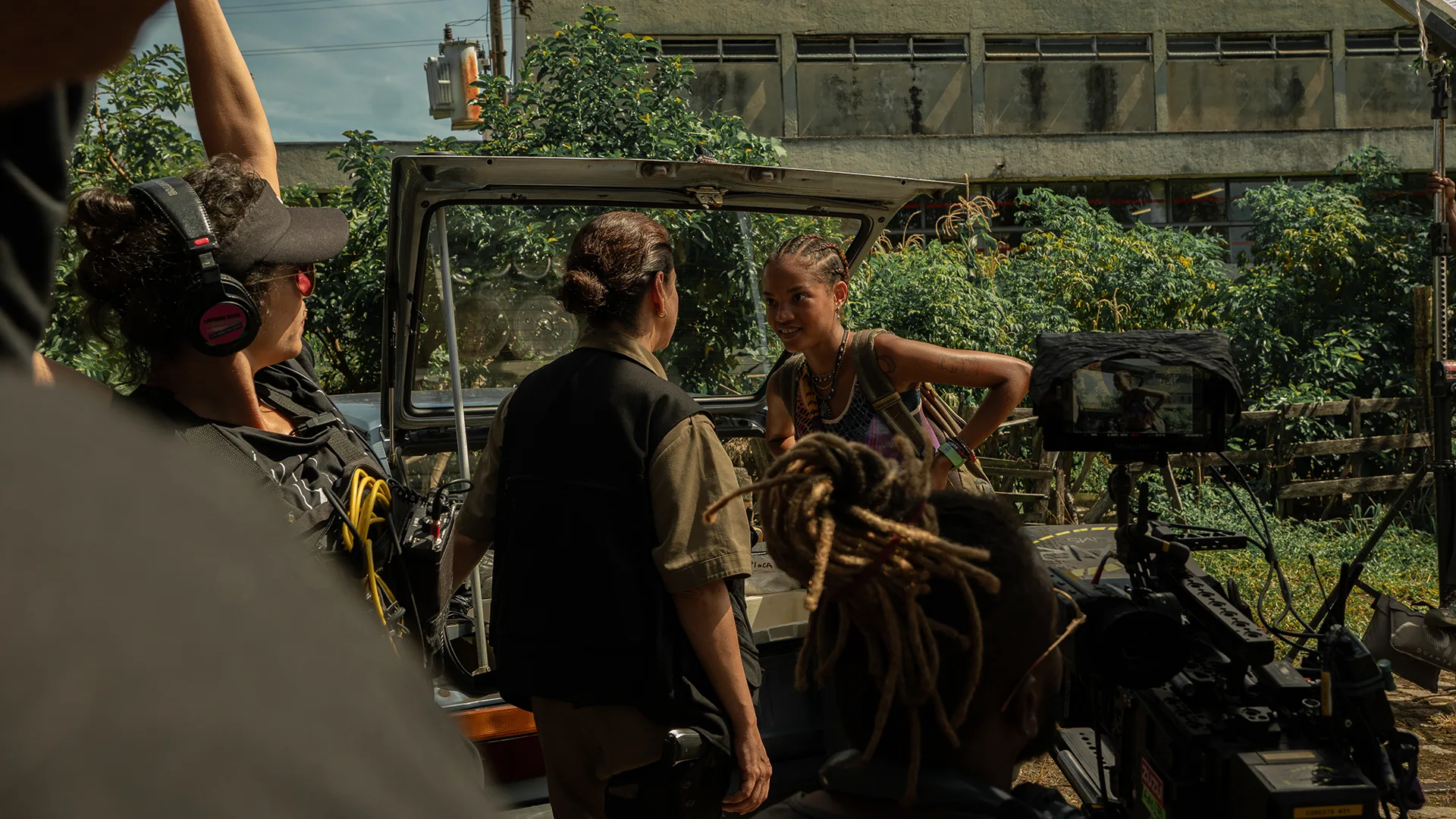 A film crew records two people talking near an open car trunk outdoors. One person wears headphones and a vest, another stands with their back to the camera, and a third smiles, leaning on the car. A camera and greenery are visible.