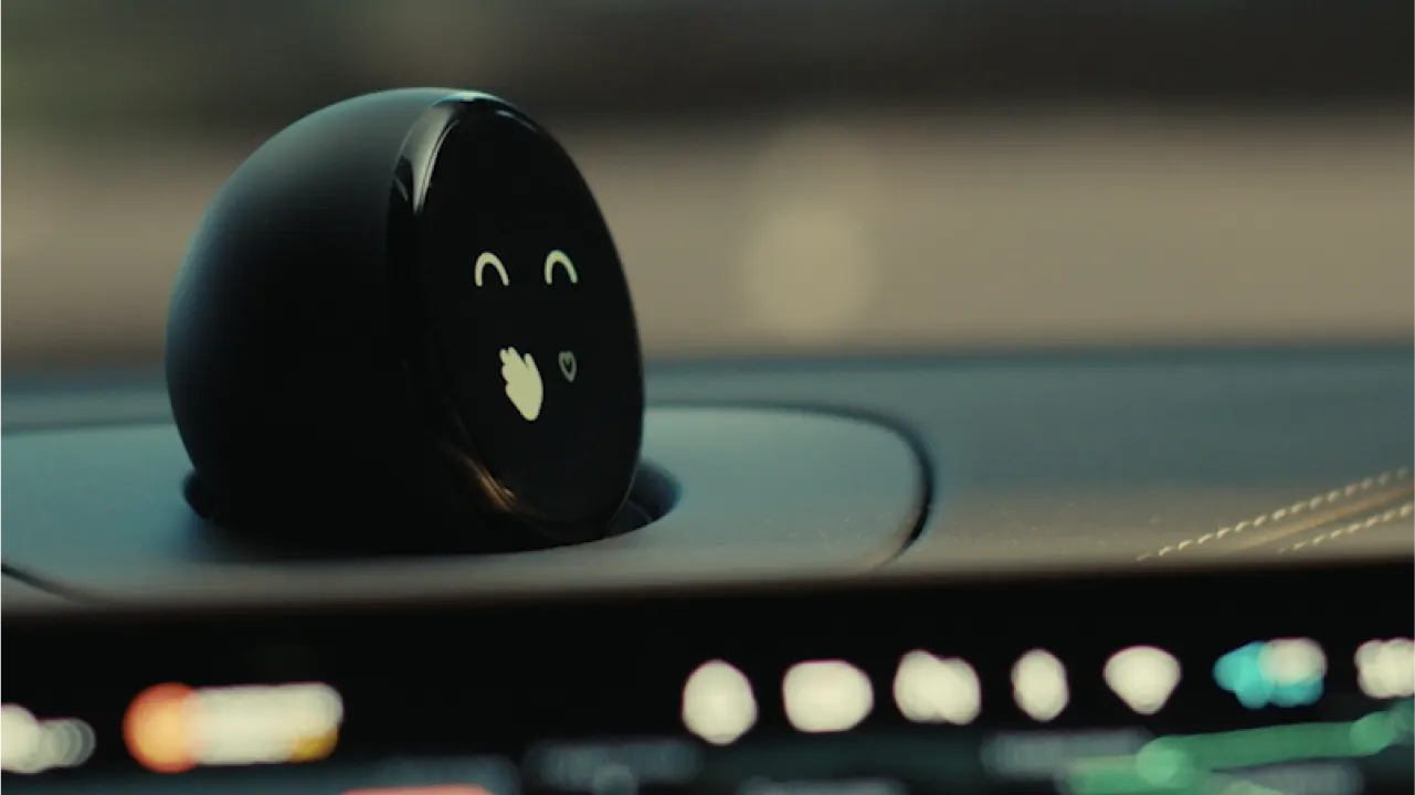 A round black digital device with a display showing a smiling face and a waving hand icon, positioned on a car dashboard. The background is blurred, highlighting the device.