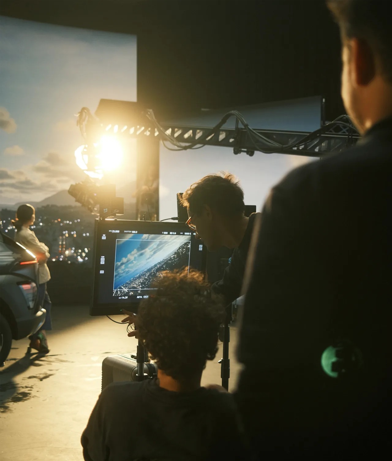 A film crew records a scene with a car and cityscape backdrop. One person monitors the footage on a screen, while others set up equipment in a studio with dramatic lighting.