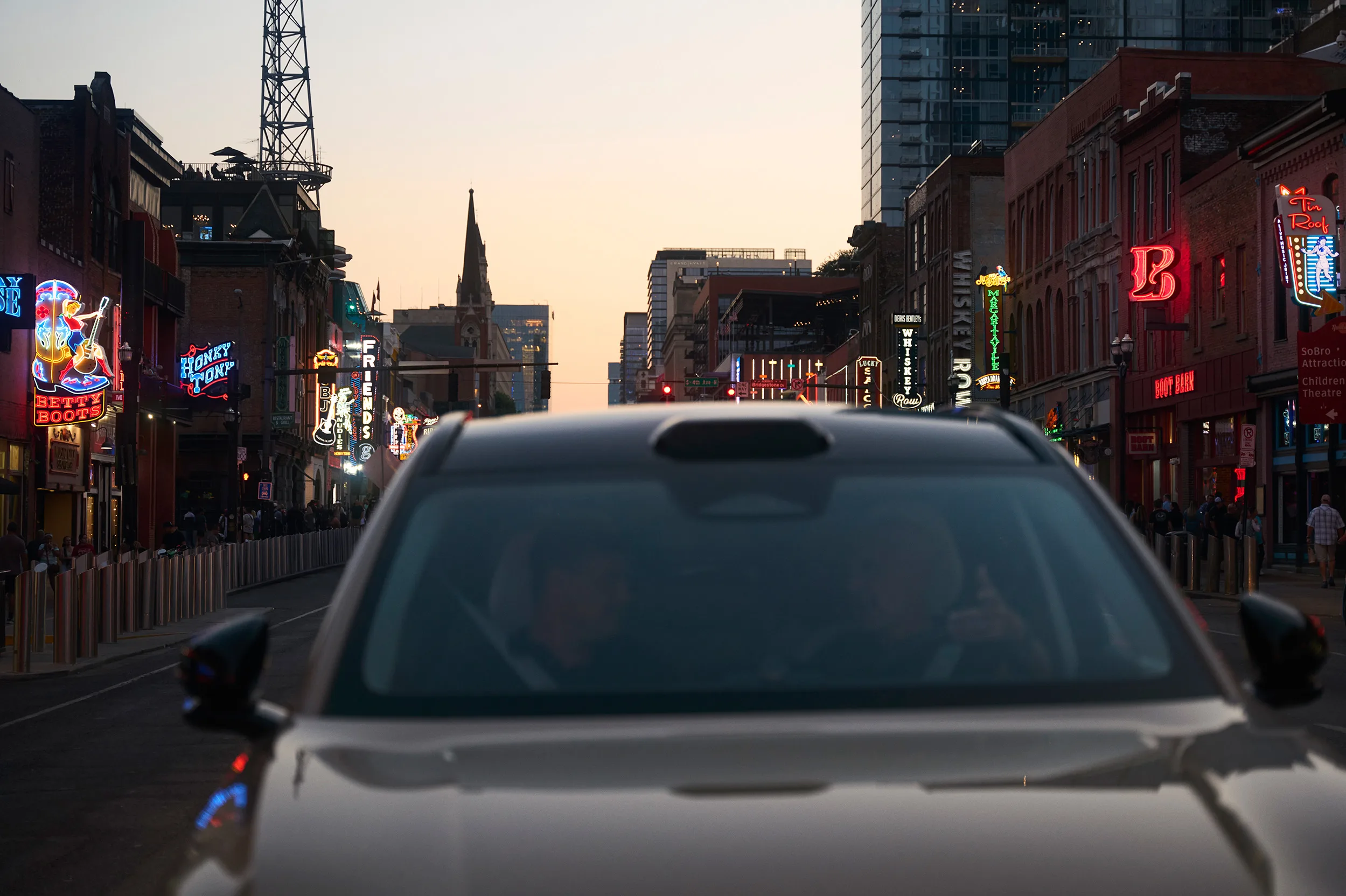 A car in the foreground with two people inside, driving down a lively city street lined with colorful neon signs and tall buildings at sunset. Crowds walk along both sides of the street.
